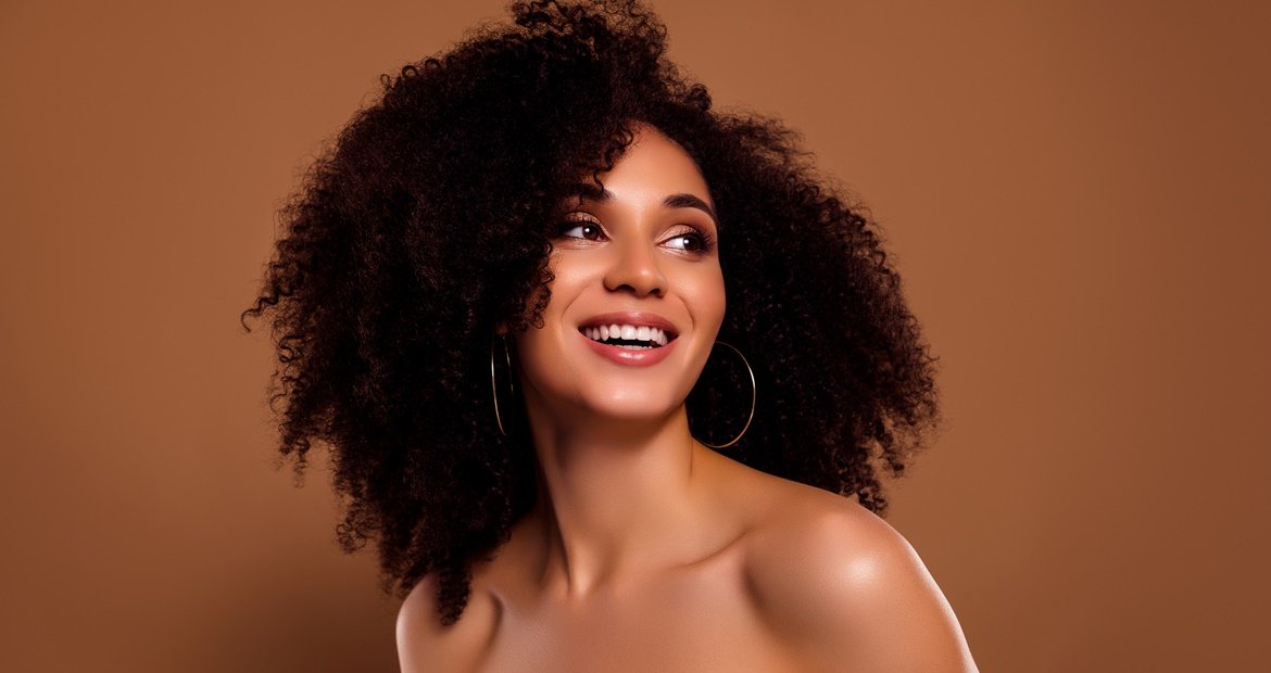 Mulher sorridente com cabelo cacheado castanho escuro olhando para o lado em fundo marrom.
